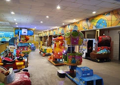 A nostalgic indoor play area featuring colorful coin-operated kiddie rides, arcade machines, and a brightly painted mural wall.