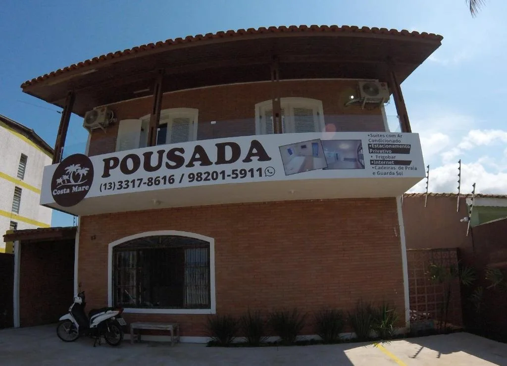 Front facade of Pousada Costa Mare brick building with large signage displaying contact information, amenities, and a parked motorcycle in the driveway.