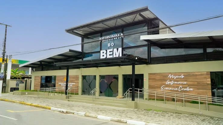 A daytime street-level view of the Feirinha do Bem building, featuring a glass upper level, green lower walls, wooden panels, and accessibility ramps.
