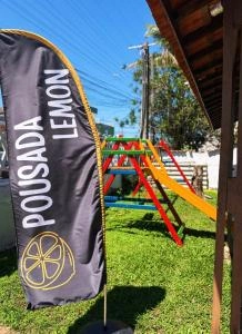 A black vertical feather flag with the words "POUSADA LEMON" and a lemon logo on a grassy lawn with a playground in the background.
