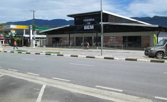 A wide street-level view of the Feirinha do Bem building under a blue sky, showing the main entrance, accessibility ramps, and a gas station in the background.