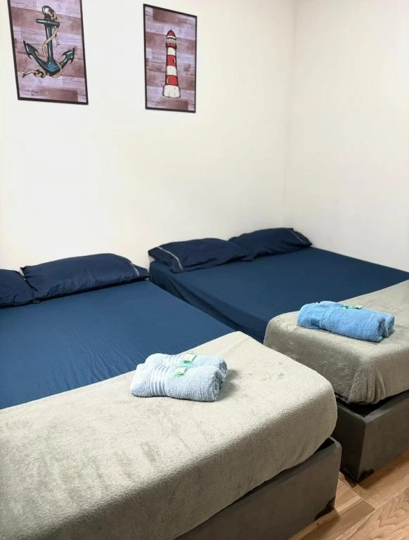 A clean bedroom featuring two beds with navy blue sheets, gray bed runners, rolled towels, and framed pictures of an anchor and lighthouse.