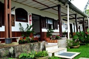 Exterior view of a tropical bungalow with a covered veranda, red brick steps, potted plants, and a lush green lawn.