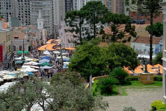 An elevated view looking down over a bustling outdoor street market with a sea of colorful umbrellas, surrounded by buildings and lush green trees.