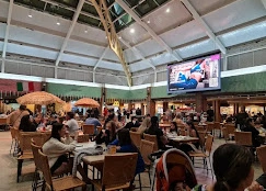 A lively and crowded food court inside a large mall with a high arched ceiling, large digital screens, and families dining at various tables.