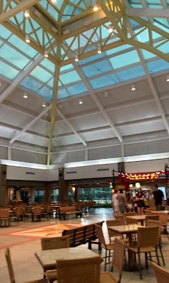 An expansive cafe seating area located under a massive, bright glass and steel skylight, featuring rattan chairs and open space.