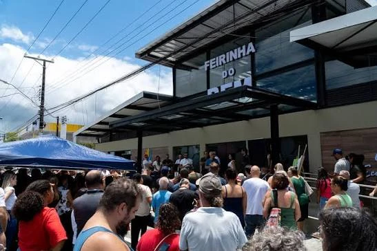A bustling outdoor scene showing a large crowd of people gathered in front of the Feirinha do Bem building, with a blue umbrella and street vendors nearby.