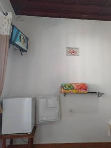 Corner of a guest room at Pousada Recanto Maresias showing a mini-fridge, wall-mounted TV, and shelf with towels.